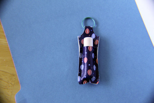 A small fabric keychain chapstick holder with a hot air balloon pattern in red and blue on a dark background, featuring a blue metal ring.